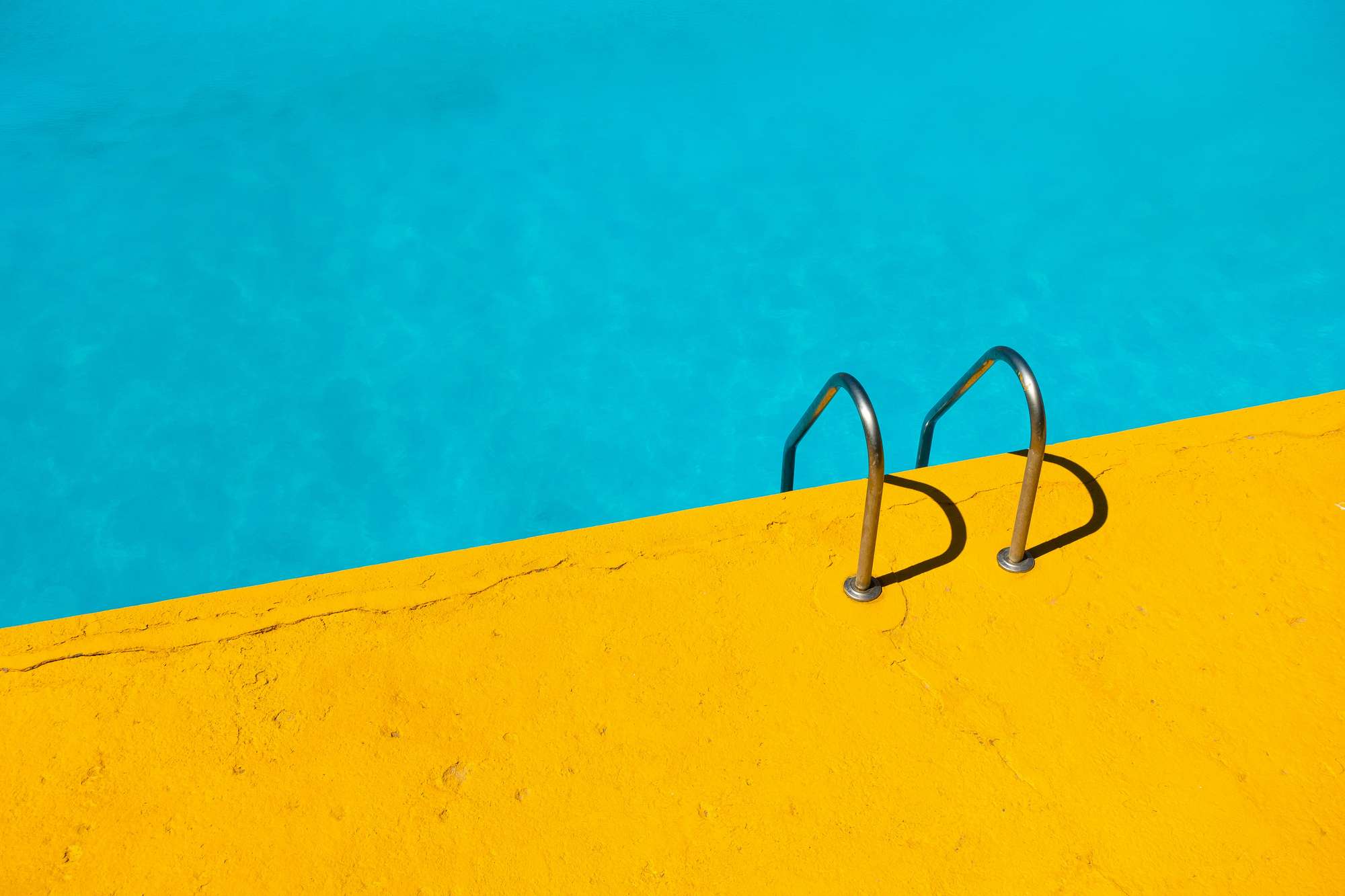 A swimming pool's edge with a stainless steel ladder; the water is a nice turquoise and the concrete around it is bright yellow