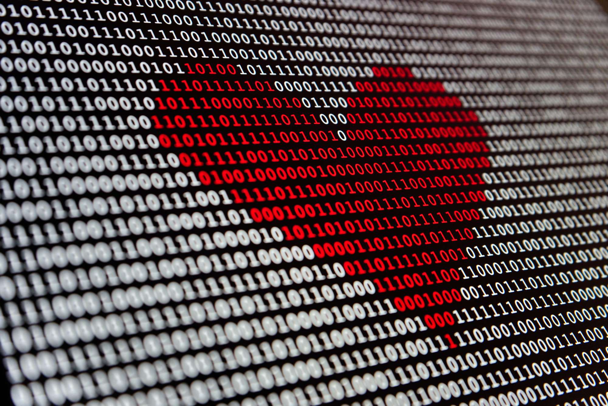 An angled photo of a surface (a screen?) that shows white and red 0s and 1s on black with the digits strategically colored so the red ones form a heart